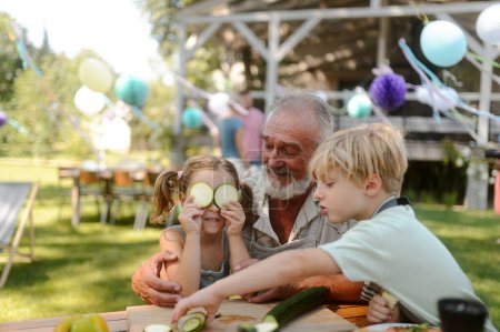 Photo for Grandfather and grandchildren having fun at a garden barbecue. Family gathering at summer garden party. Granddauhter putting cucumber slices on her eyes. - Royalty Free Image