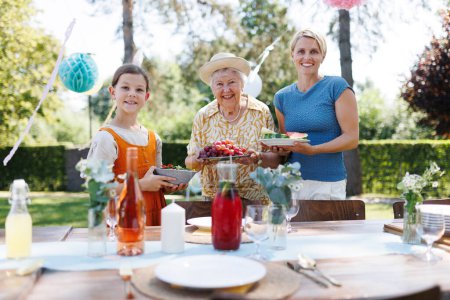 Photo for Grandmother, mother and daughter setting table for summer garden party. Three generations of women in family, bringing plates, food, and drinks at table. - Royalty Free Image