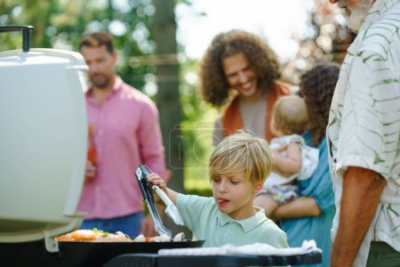 Téléchargez les photos : Jeune garçon tournant la nourriture grillée avec des pinces. Grand-père et petit-fils grillent ensemble à une fête barbecue jardin. - en image libre de droit