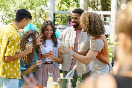 Téléchargez les photos : Les amis et la famille parlent et s'amusent lors d'une fête de jardin sur le gril d'été. Les gens à la fête riant, tenant des verres avec des boissons. - en image libre de droit