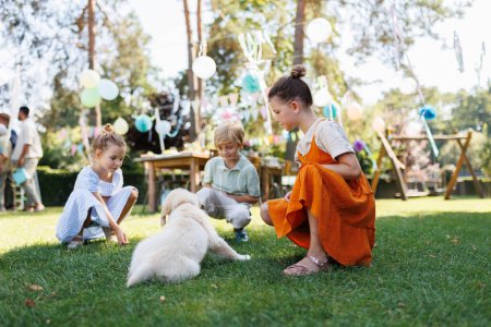 Foto de Niños jugando con un cachorro pequeño en una fiesta de jardín familiar. Reunión familiar al aire libre durante el cálido día de otoño o verano. - Imagen libre de derechos