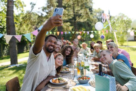 The whole family sitting at the table taking a group selfie at the family garden party. A family gathering to celebrate a birthday.