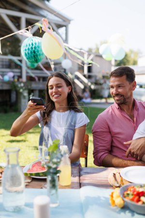 Téléchargez les photos : Femme prenant une photo de sa nourriture avec son smartphone lors d'une garden party. Family bbq garden party. - en image libre de droit