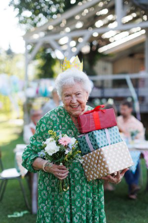 Foto de Fiesta de cumpleaños en el jardín para la señora mayor. Hermosa mujer de cumpleaños con corona de papel en la cabeza y regalos en las manos. - Imagen libre de derechos