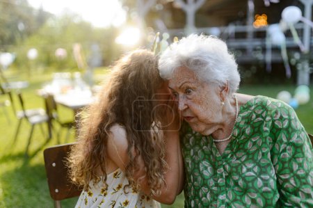 Photo for Young girl whispering a secret into grandmothers ear at a garden party. Love and closeness between grandparent and grandchild. - Royalty Free Image