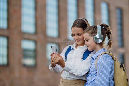 Foto de Dos colegialas están revisando las redes sociales en su teléfono inteligente frente a la escuela. Amigos con auriculares están viendo un video. - Imagen libre de derechos