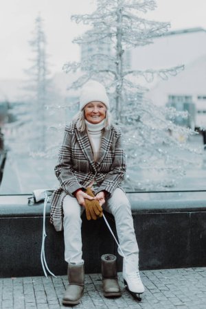 Téléchargez les photos : Femme âgée assise au rinçage de glace de Noël dans la ville et mise sur des patins à glace blancs. Apprendre à patiner dans la vieillesse. - en image libre de droit