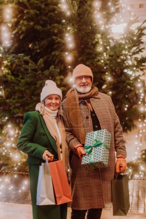 Foto de Feliz pareja de ancianos disfrutando del mercado de Navidad al aire libre en la noche, la compra de regalos y árbol de Navidad. - Imagen libre de derechos