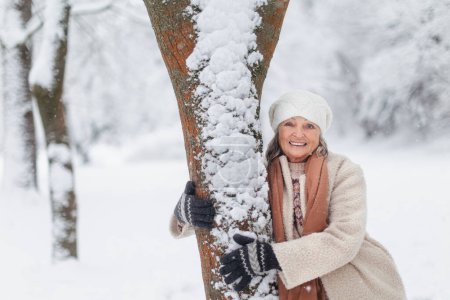 Téléchargez les photos : Portrait taille haute d'une élégante femme âgée debout près d'un arbre, pendant la froide journée enneigée d'hiver. Femme âgée passant des vacances d'hiver dans les montagnes. Paysage hivernal de Noël. - en image libre de droit