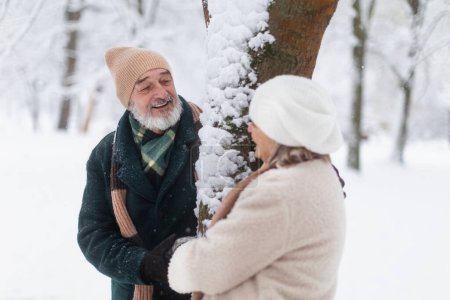 Téléchargez les photos : Élégant couple de personnes âgées debout près d'un arbre dans le parc enneigé, pendant la froide journée enneigée d'hiver. Couple de personnes âgées passant des vacances d'hiver dans les montagnes. Paysage hivernal de Noël. - en image libre de droit