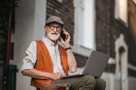 Senior man sitting on street curb working on his laptop outdoors. Portrait of elderly man using digital technologies, working with notebook and smartphone. Concept of seniors and digital skills.