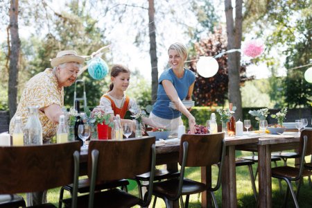 Photo for Grandmother, mother and daughter setting table for summer garden party. Three generations of women in family, bringing plates, food and drinks at party table. Getting ready for family gathering. - Royalty Free Image
