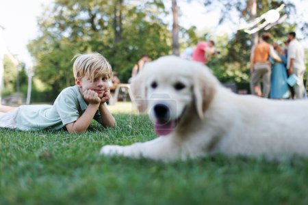 Foto de Niños jugando con un cachorro pequeño en una fiesta de jardín familiar. Reunión familiar al aire libre durante el cálido día de otoño. Retrato de niño tendido en la hierba con Golden retriever cachorro. - Imagen libre de derechos