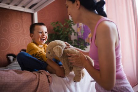 Photo for A single mother waking her son up for school in the morning. Beautiful mom awakens the schoolboy with a stuffed toy, playing and making him laugh. - Royalty Free Image