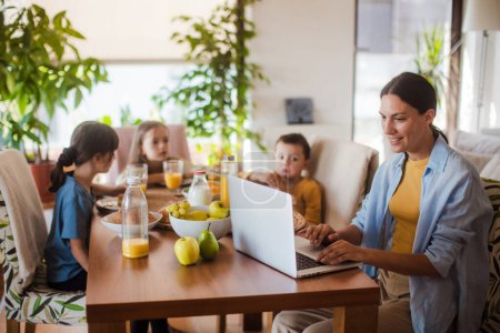 Foto de Madre trabajando en un portátil en la mesa de la cocina mientras los niños desayunan. Oficina de trabajo remoto y hogar para madres con niños. - Imagen libre de derechos