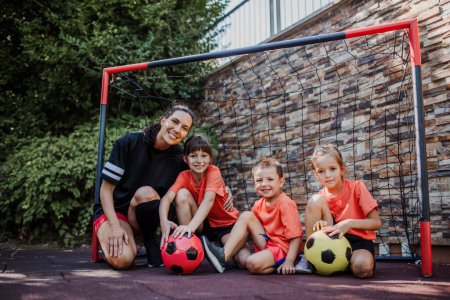 Photo for Mom playing football with her children, dressed in football jerseys. The family as one soccer team. Fun family sports activities outside in the backyard or on the street. - Royalty Free Image