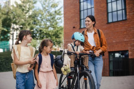 Photo for Mom picking up daughters from school with little son in bike seat . The schoolgirls telling mother about their day at school, walking in front of the school building. - Royalty Free Image