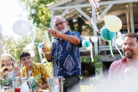 Photo for Grandfather opening a bottle of champagne, pouring a champagne into glasses. Senior man making a celebratory toast at an outdoor summer garden party. - Royalty Free Image