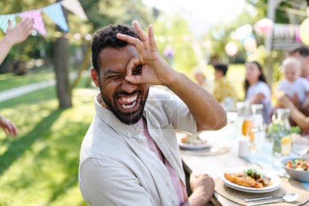Photo for Portrait of handsome man making funny faces, sitting at the party table during a summer garden party with family and friends. - Royalty Free Image