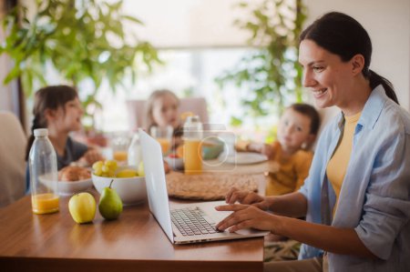 Téléchargez les photos : Mère travaillant sur un ordinateur portable à la table de la cuisine pendant que les enfants prennent le petit déjeuner. Travail à distance et bureau à domicile pour les mères avec enfants. - en image libre de droit