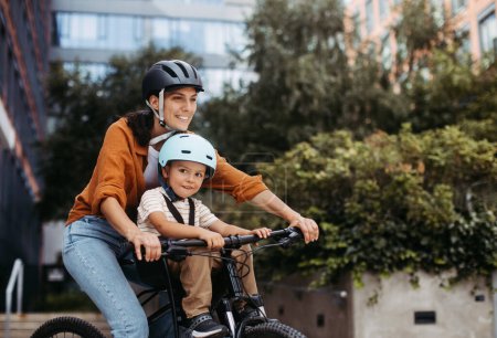 Foto de Madre llevando a su hijo en un seguro portabicicletas o asiento, ambos con cascos. Mamá viajando con un niño pequeño a través de la ciudad en bicicleta. - Imagen libre de derechos