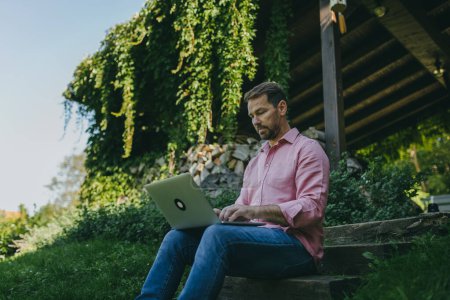 Photo for Low angle shot of man working outdoors in the garden, with laptop on his legs. Businessman working remotely from homeoffice, thinking about new business or creative idea. - Royalty Free Image