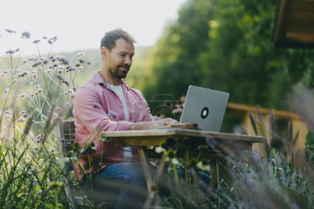 Photo for Low angle shot of man working outdoors in the garden, with laptop on his legs. Businessman working remotely from homeoffice, thinking about new business or creative idea. - Royalty Free Image