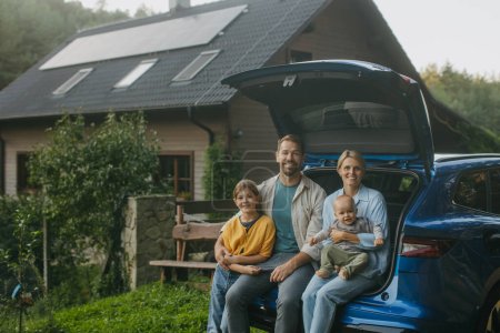 Téléchargez les photos : Famille assise dans le coffre de la voiture électrique, avec maison avec des panneaux solaires sur le toit derrière eux. Énergie solaire et mode de vie durable de la jeune famille. Concept d'énergie verte et avenir durable - en image libre de droit
