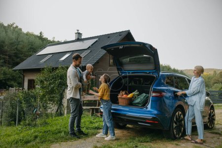 Foto de Familia con coche eléctrico de pie en frente de su casa con paneles solares en el techo. Energía solar y estilo de vida sostenible de la familia joven. Concepto de energía verde y futuro sostenible para el próximo - Imagen libre de derechos