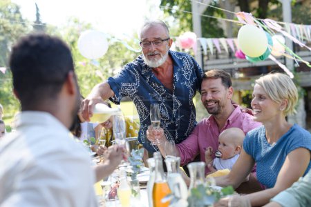 Grandfather opening a bottle of champagne, pouring a champagne into glasses. Senior man making a celebratory toast at an outdoor summer garden party.