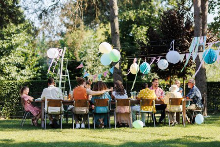 Photo for Family and friends sitting at the party table during a summer garden party outdoors. Rear view of people sitting at the table outdoors. - Royalty Free Image