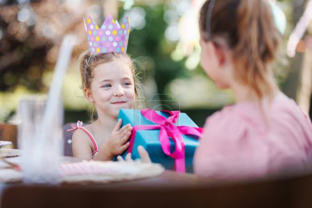 Little friends at a birthday party outdoors at garden. Cute girl with paper crown giving the birthday girl gift. Birthday garden party for children.