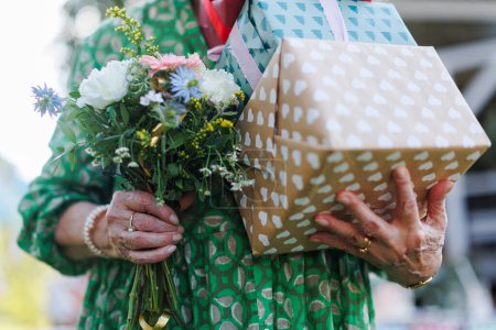 Téléchargez les photos : Gros plan de la femme d'anniversaire senior tenant des cadeaux et un bouquet dans les mains. Fête d'anniversaire de jardin pour dame senior. - en image libre de droit