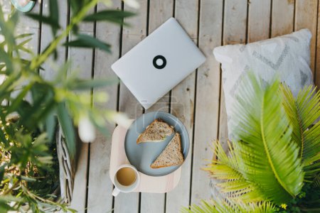 Téléchargez les photos : Vue de dessus de jardin extérieur patio avec ordinateur portable, assiette avec déjeuner léger et tasse de café. Cadre de bureau à domicile extérieur simple pour employé à distance, télécommuter. - en image libre de droit