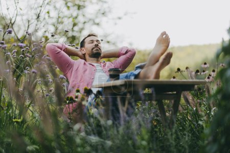 Téléchargez les photos : Homme pieds nus détendu assis dans le jardin avec les pieds sur la table. Père ayant un moment avec lui-même pendant que ses enfants sont à l'école. - en image libre de droit