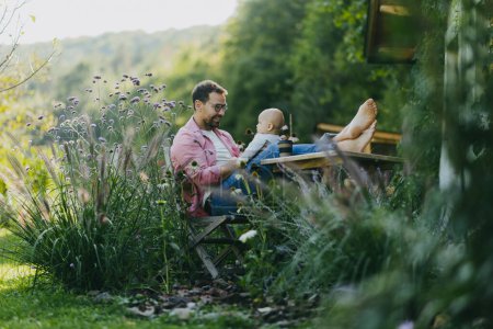Foto de Hombre sentado en el jardín con los pies en la mesa, jugando con su hijo pequeño. Padre teniendo un momento de unión con su hijo pequeño al aire libre. - Imagen libre de derechos