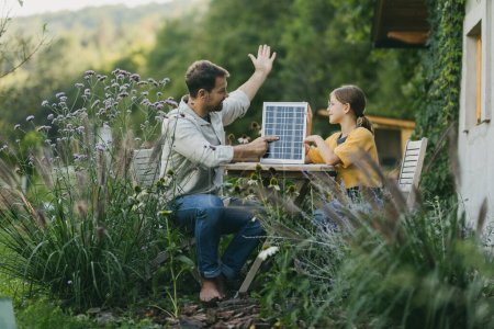 Téléchargez les photos : Père avec sa fille assise à l'extérieur dans le jardin, avec modèle de panneau solaire. Énergie solaire et mode de vie durable de la jeune famille. Concept d'énergie verte et d'avenir durable pour la prochaine - en image libre de droit