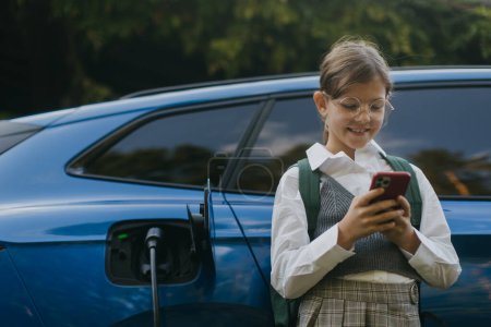Photo for Girl and electric car with charger in charging port. Girl scrolling on her smartphone while care is charging before going to school. - Royalty Free Image