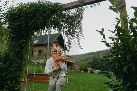 Foto de Hermosa madre y el bebé balanceándose en un columpio en el jardín, divertirse y pasar tiempo de calidad juntos al aire libre durante un cálido día de otoño. - Imagen libre de derechos