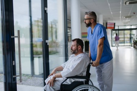 Male nurse pushing a patient in wheelchair along a hospital corridor. Overweight patient feeling anxious and has health concerns.