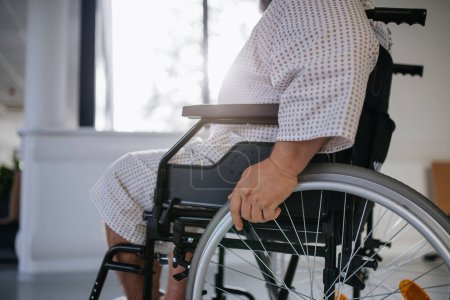 Téléchargez les photos : Vue latérale de l'homme en fauteuil roulant. Patient en surpoids en robe d'hôpital en attente d'un examen médical à l'hôpital. - en image libre de droit