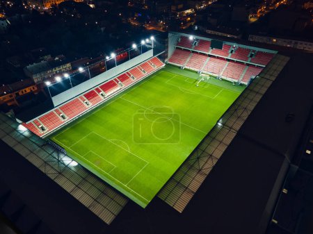 Téléchargez les photos : Vue aérienne d'un stade de football illuminé avec un toit ouvert la nuit. Terrain de football avec stands vides en ville. - en image libre de droit