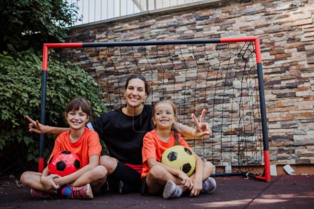 Photo for Mom playing football with her daughters, dressed in football jerseys. The family as one soccer team. Fun family sports activities outside in the backyard or on the street. - Royalty Free Image