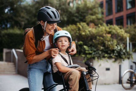 Photo for Mother fastening sons bike helmet on head, carring him on child bike carrier or seat. Mom commuting with a young child through the city on a bicycle. - Royalty Free Image
