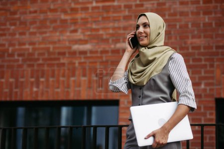 Téléchargez les photos : Portrait de belle femme en hijab debout sur la rue de la ville. Femme d'affaires musulmane avec ordinateur portable passer un appel. Iran, Afganistan enseignante devant le bâtiment de l'école. - en image libre de droit