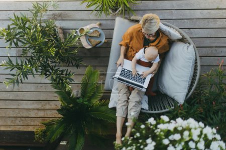 Photo for Top view of mother holding baby while working on laptop in garden. Businesswoman working remotely from outdoor home office and taking care of little son. Life work balance with kid. Working mom with - Royalty Free Image