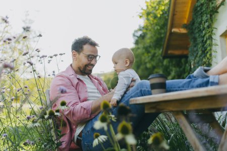 Photo for Man sitting in the garden with feet on table, playing with his baby son. Father having bonding moment with his little cute kid. - Royalty Free Image