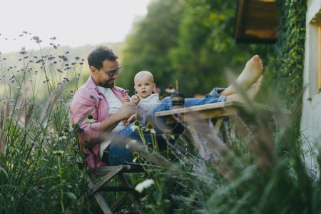 Téléchargez les photos : Homme assis dans le jardin avec les pieds sur la table, jouant avec son bébé fils. Mon père s'est lié d'amitié avec son petit mignon. Bébé jouer avec la fleur. - en image libre de droit