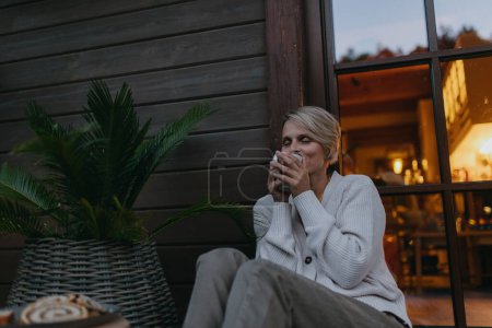 Téléchargez les photos : Angle bas de la femme assise sur la terrasse pendant le crépuscule d'automne, sirotant du thé chaud et profitant de la vue. À l'intérieur de la maison derrière elle, son chaud et accueillant, alors qu'elle est habillée d'un pull confortable - en image libre de droit