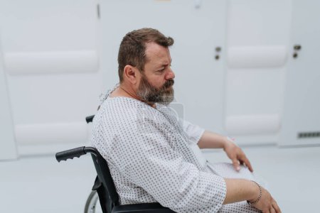 Foto de Vista lateral del hombre en silla de ruedas. Paciente con sobrepeso en bata de hospital esperando un examen médico en el hospital, sintiéndose ansioso. - Imagen libre de derechos